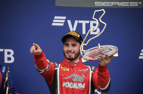 Joey Mawson on the podium in a red race suit holding a trophy